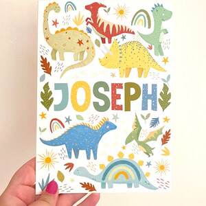 以下が含まれることがあります： 白い背景に、さまざまな恐竜と「JOSEPH」という名前が大きなカラフルな文字で書かれたカラフルなプリント。恐竜は緑、青、黄、赤、オレンジの色合いです。