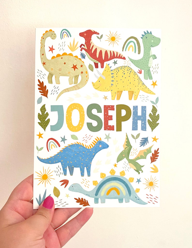 以下が含まれることがあります： 白い背景に、さまざまな恐竜と「JOSEPH」という名前が大きなカラフルな文字で書かれたカラフルなプリント。恐竜は緑、青、黄、赤、オレンジの色合いです。