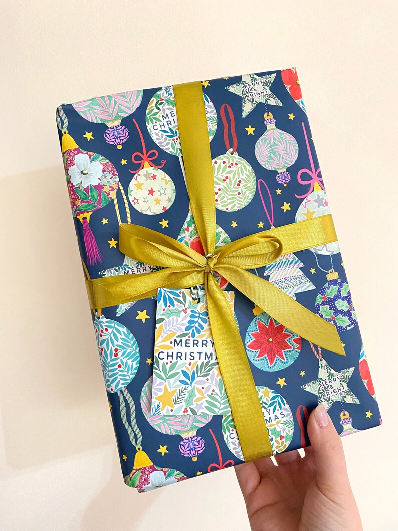 May include: A blue gift wrap with a gold ribbon and a gift tag that says "Merry Christmas". The gift wrap has a pattern of colorful ornaments and stars.