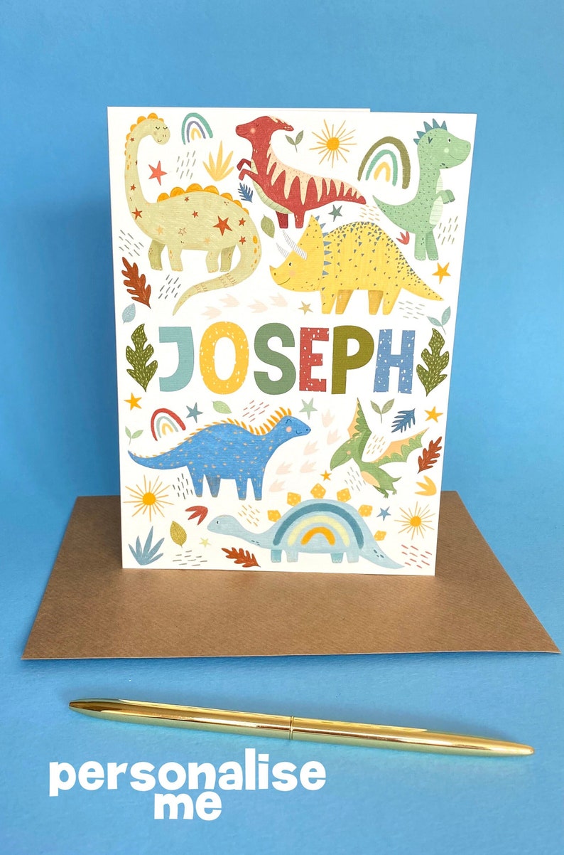 以下が含まれることがあります： カラフルな恐竜のデザインと「JOSEPH」という名前が大きなカラフルな文字で書かれた白いカード。カードは茶色の封筒と金のペンで囲まれています。