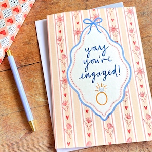 Engagement greeting card shown on wooden backdrop. Card reads yay you're engaged inside of a blue ribbon frame with florals surrounding