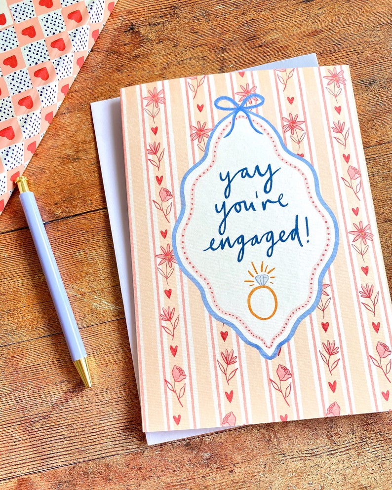Engagement greeting card shown on wooden backdrop. Card reads yay you're engaged inside of a blue ribbon frame with florals surrounding