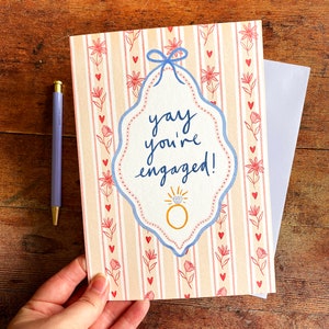 Engagement greeting card shown on wooden backdrop. Card reads yay you're engaged inside of a blue ribbon frame with florals surrounding