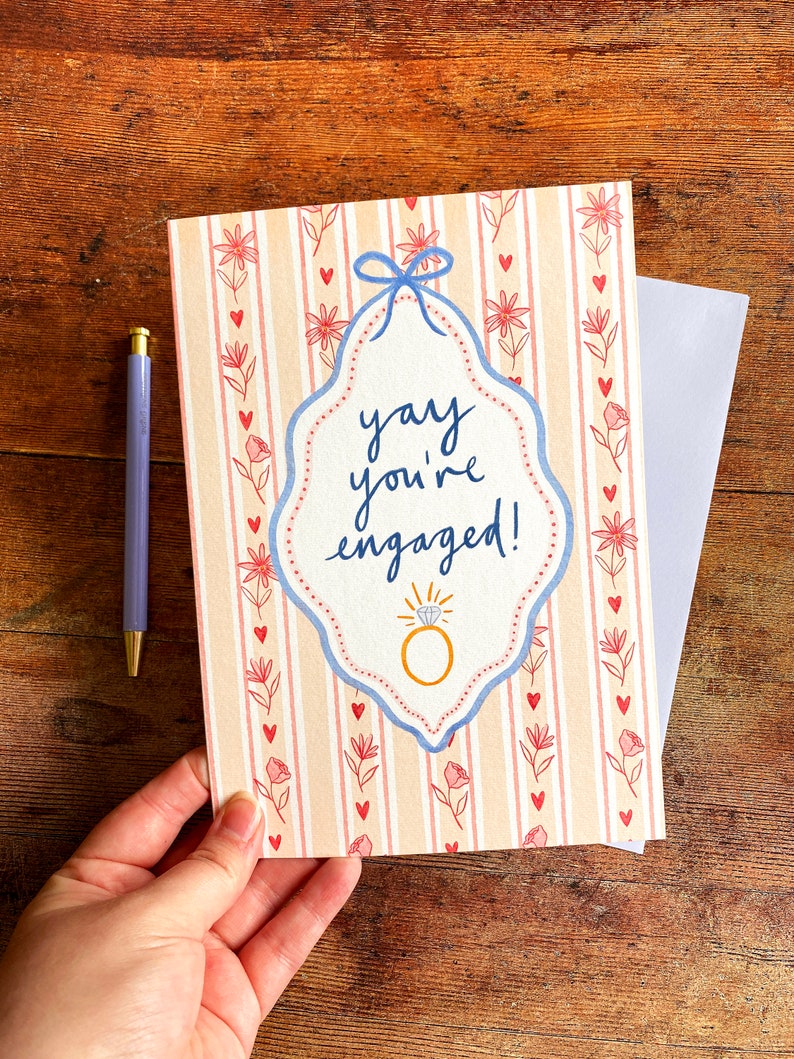 Engagement greeting card shown on wooden backdrop. Card reads yay you're engaged inside of a blue ribbon frame with florals surrounding