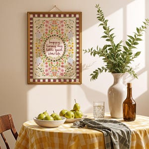 May include: A rectangular wall hanging in a gold frame featuring a floral border with birds and the text "happy living my little quiet slow life" in the center, displayed above a table with a yellow checkered cloth, pears, and a vase.