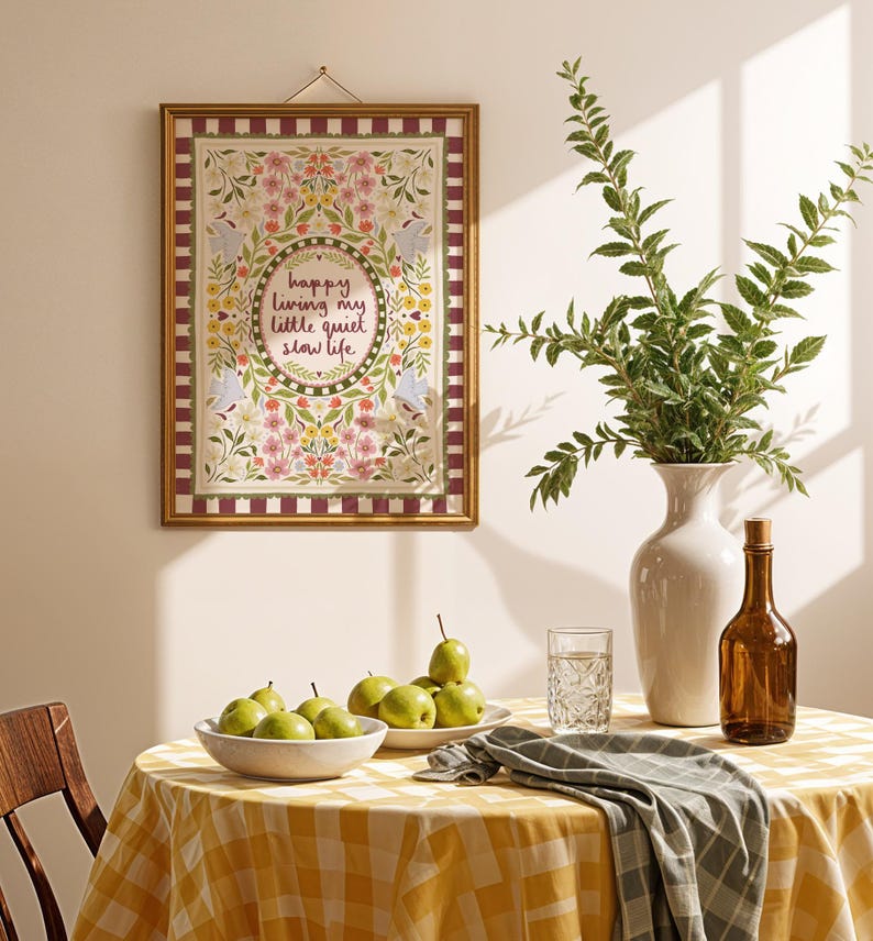 May include: A rectangular wall hanging in a gold frame featuring a floral border with birds and the text "happy living my little quiet slow life" in the center, displayed above a table with a yellow checkered cloth, pears, and a vase.