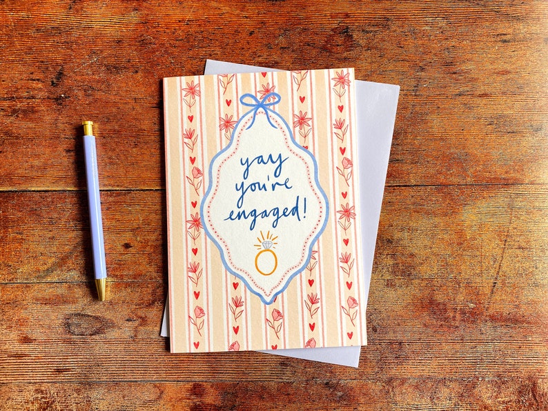 Engagement greeting card shown on wooden backdrop. Card reads yay you're engaged inside of a blue ribbon frame with florals surrounding