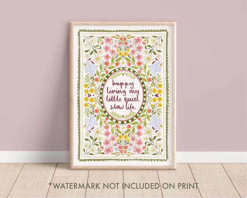 May include: A framed art print with a floral design and the text "happy living my little quiet slow life." The artwork features a white background with pink, yellow, and green floral elements, and two blue bird illustrations. The frame is a light wood colour.