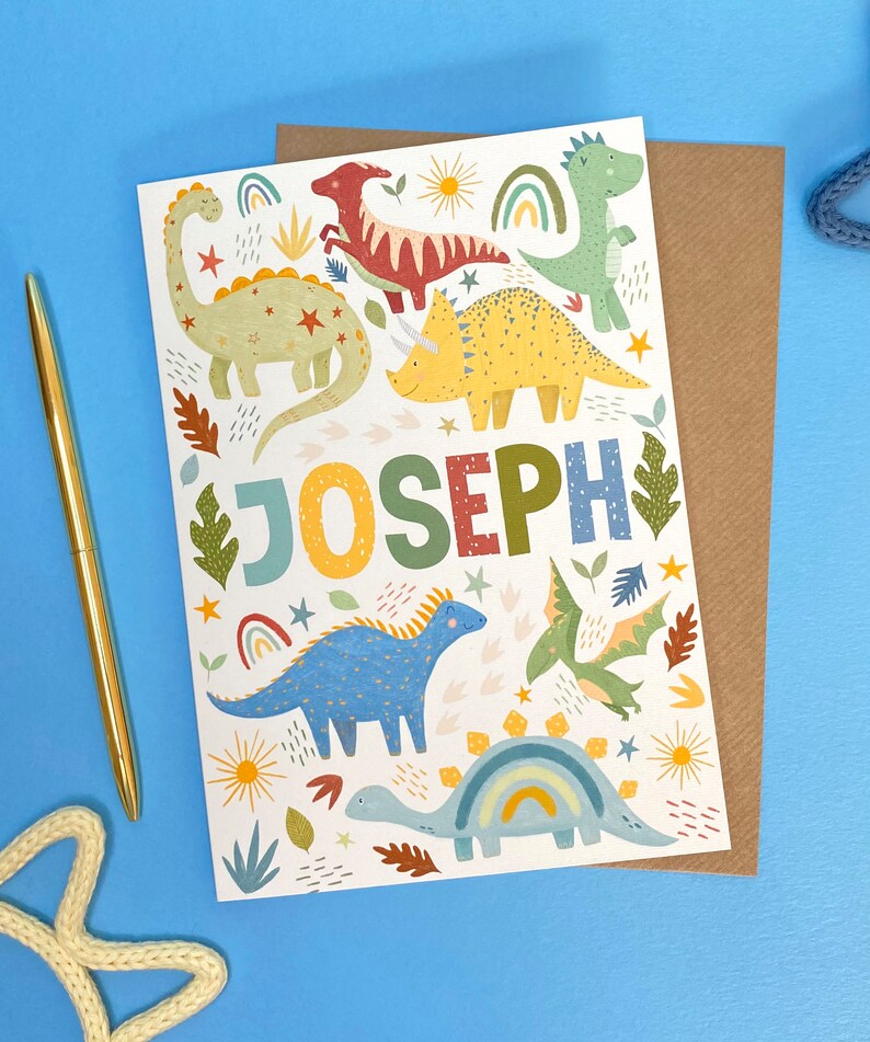以下が含まれることがあります： カラフルな恐竜をテーマにした白いカード。カードには、緑色の恐竜、赤色の恐竜、黄色の恐竜、青色の恐竜、緑と青の恐竜など、さまざまな色の恐竜がさまざまなパターンで描かれています。カードには、カラフルな文字で「JOSEPH」という名前も書かれています。