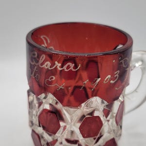 May include: A red glass mug with a clear, faceted design. The mug has a handle and is inscribed with the text "Clara to Et. 1903".