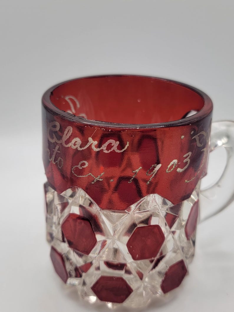 May include: A red glass mug with a clear, faceted design. The mug has a handle and is inscribed with the text "Clara to Et. 1903".