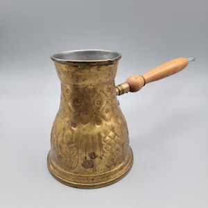 May include: A vintage brass coffee pot with a wooden handle. The pot has an ornate, embossed design and a silver-colored interior. The handle is made of light-colored wood and is attached to the side of the pot. The pot is designed for brewing coffee.