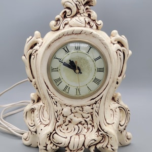 May include: A white ceramic clock with ornate details and Roman numeral markings on the face. The clock has a white cord and is plugged in.