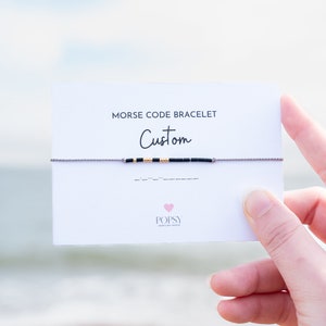 May include: A white card with the text "Morse Code Bracelet Custom" and a grey string bracelet with black and gold beads. The bracelet is spelled out in Morse code on the card. The card also has the logo "POPSY Jewellery Design" with a pink heart.