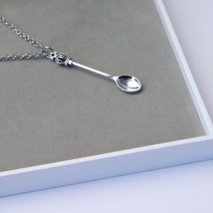 May include: A silver spoon necklace with a crown-shaped pendant. The spoon is hanging on a silver chain and is displayed on a grey background.