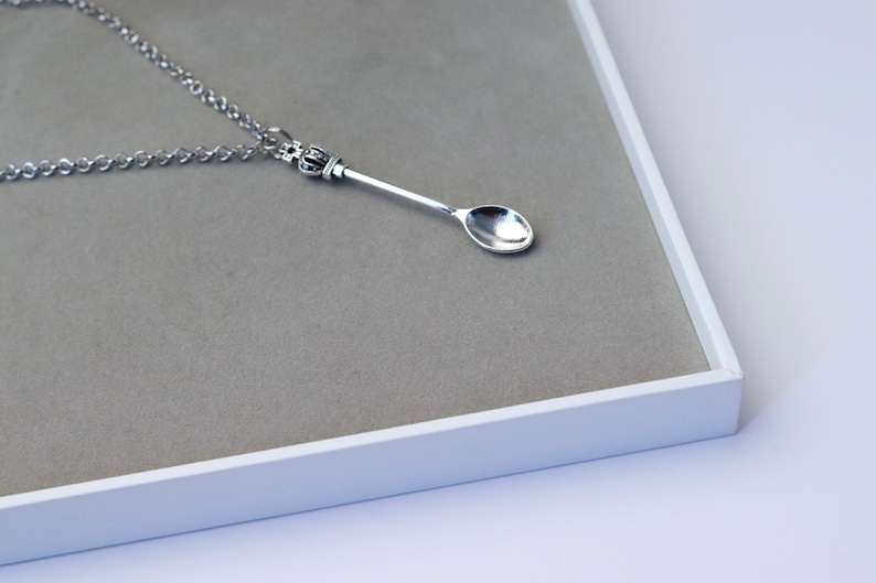 May include: A silver spoon necklace with a crown-shaped pendant. The spoon is hanging on a silver chain and is displayed on a grey background.