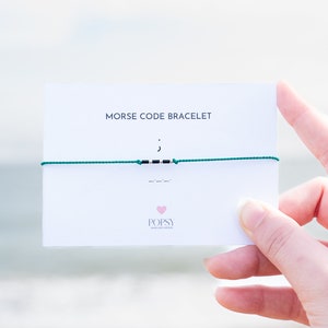 May include: A white card with the text "Morse Code Bracelet" and a green string bracelet with black beads spelling out the Morse code for the letter "J". The card also has the logo "POPSY Jewelry & Gifts" with a pink heart.