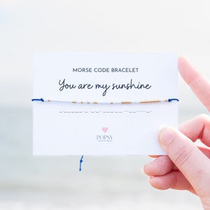 May include: White beaded bracelet with gold beads spelling out "You are my sunshine" in Morse code on a white card with the text "Morse Code Bracelet" and the brand name "POPSY".