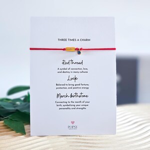 May include: A red string bracelet with a gold charm that says "LUCKY" and a blue gemstone. The bracelet is on a white card with the text "THREE TIMES A CHARM", "Red Thread", "A symbol of connection, love, and destiny in many cultures", "Luck", "Believed to bring good fortune, protection, and positive energy", "March birthstone", "Connecting to the month of your birth, symbolizing your unique personality and strengths", and "POPSY".