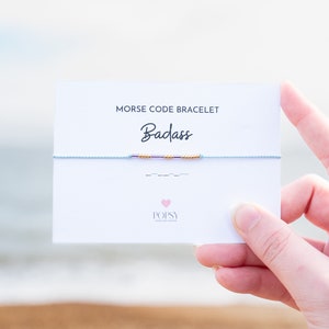 May include: A white card with the text "MORSE CODE BRACELET" and "Badass" in black. A blue string bracelet with gold and purple beads is displayed on the card. The card also has the text "POPSY" in pink with a pink heart.