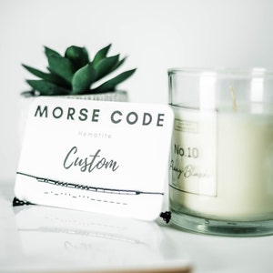 May include: A white card with the text "Morse Code" and "Hematite" printed on it. The card also has the word "Custom" written in cursive. A black string bracelet with silver beads is displayed on the card. The bracelet is made of hematite beads.