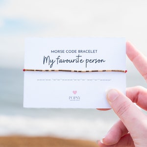 May include: A white card with the text "Morse Code Bracelet My favourite person" and a red and gold beaded bracelet. The card also has the text "POPSY Jewellery Design" and a pink heart.