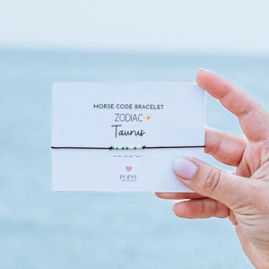 May include: A brown string bracelet with green and silver beads spelling out "Taurus" in Morse code. The bracelet is on a white card with the text "Morse Code Bracelet Zodiac Taurus".