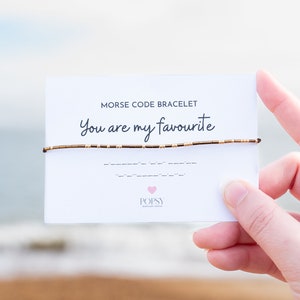 Peut inclure: Une carte blanche avec le texte "Bracelet en code Morse" et "You are my favourite" en noir. Un bracelet en perles brunes est enroulé autour de la carte. La carte a également un cœur rose avec le mot "POPSY" en noir.