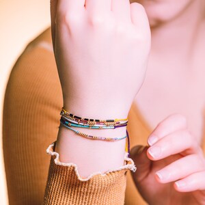 May include: A person's wrist with multiple colorful beaded bracelets. The bracelets are layered and include shades of blue, purple, yellow, and black.