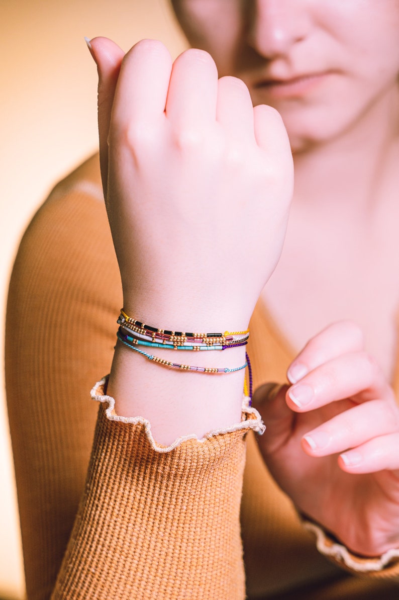 May include: A person's wrist with multiple colorful beaded bracelets. The bracelets are layered and include shades of blue, purple, yellow, and black.