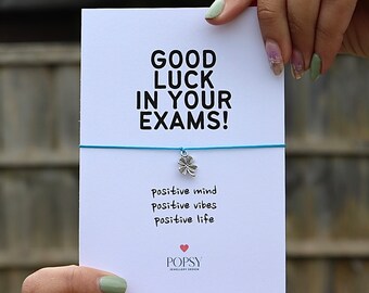 May include: A white card with the text "GOOD LUCK IN YOUR EXAMS!" and "positive mind positive vibes positive life." A silver four-leaf clover charm hangs from a blue string. The card is branded with the "POPSY JEWELLERY DESIGN" logo.
