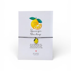 May include: A white card featuring a lemon illustration and the phrase "Squeeze the day." A black string bracelet with a lemon charm is attached. The card also has instructions and the brand name POPSY.