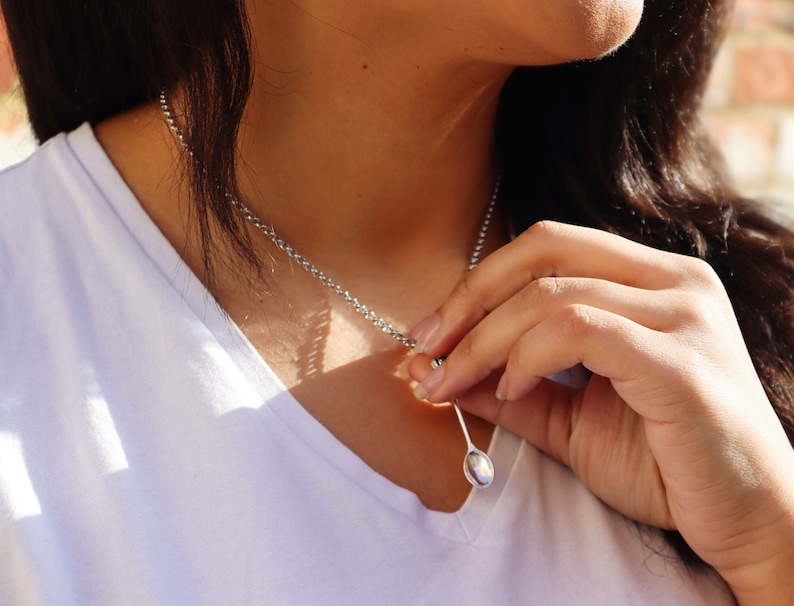 May include: A silver chain necklace with a small, oval-shaped pendant. The pendant is a spoon.