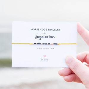 May include: A yellow string bracelet with black and gold beads spelling out "Vegetarian" in Morse code. The bracelet is on a white card with the text "Morse Code Bracelet Vegetarian" and the brand name "POPSY Jewelry Design".