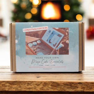 May include: A cardboard box kit labeled "Christmas Edition" with the text "Make Your Own Morse Code Bracelets." The box features an image of the kit's contents, including beads and instructions. The background shows a blurred fireplace.
