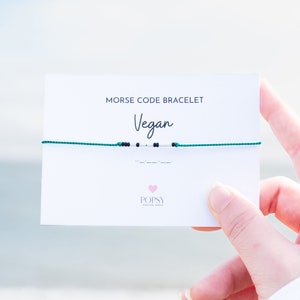 May include: A green string bracelet with white and black beads spelling out the word "Vegan" in Morse code. The bracelet is on a white card with the text "Morse Code Bracelet Vegan" and the logo "POPSY Jewelry Design".