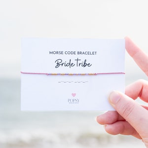 May include: A pink and gold beaded bracelet with the words "Bride Tribe" on a white card. The bracelet is a Morse code bracelet with the code for "Bride Tribe" written below the bracelet.
