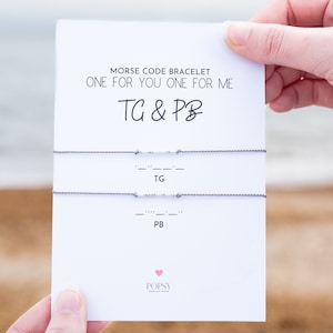 May include: A white card with the text "Morse Code Bracelet One for You One for Me TG & PB" and two grey strings with white beads representing the morse code for "TG" and "PB".