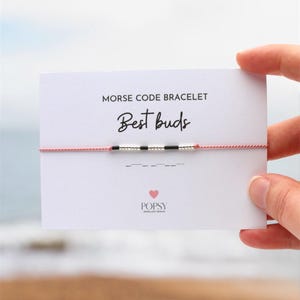 May include: A pink string bracelet with black and white beads spelling out "Best buds" in Morse code. The bracelet is on a white card with the text "Morse Code Bracelet" and "Best buds" written in black.