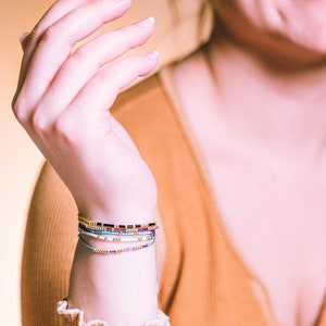 May include: A close-up of a person's wrist wearing a stack of colorful beaded bracelets. The bracelets are made of small, colorful beads and are arranged in a layered fashion. The bracelets are a mix of yellow, blue, purple, white, and black beads.
