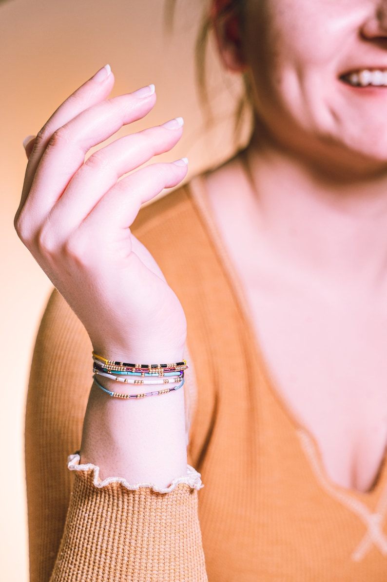 May include: A close-up of a person's wrist wearing a stack of colorful beaded bracelets. The bracelets are made of small, colorful beads and are arranged in a layered fashion. The bracelets are a mix of yellow, blue, purple, white, and black beads.