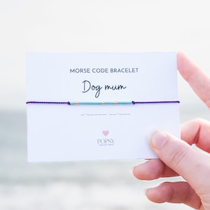 May include: A purple string bracelet with blue and gold beads spelling out "Dog mum" in Morse code. The bracelet is on a white card with the text "Morse Code Bracelet" and "Popsy Jewellery Design".