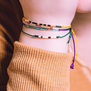 May include: A close-up of a person's wrist wearing several colorful beaded bracelets. The bracelets are made of small beads in various colors, including green, blue, yellow, purple, and black. The bracelets are tied with knots and have long, dangling ends.