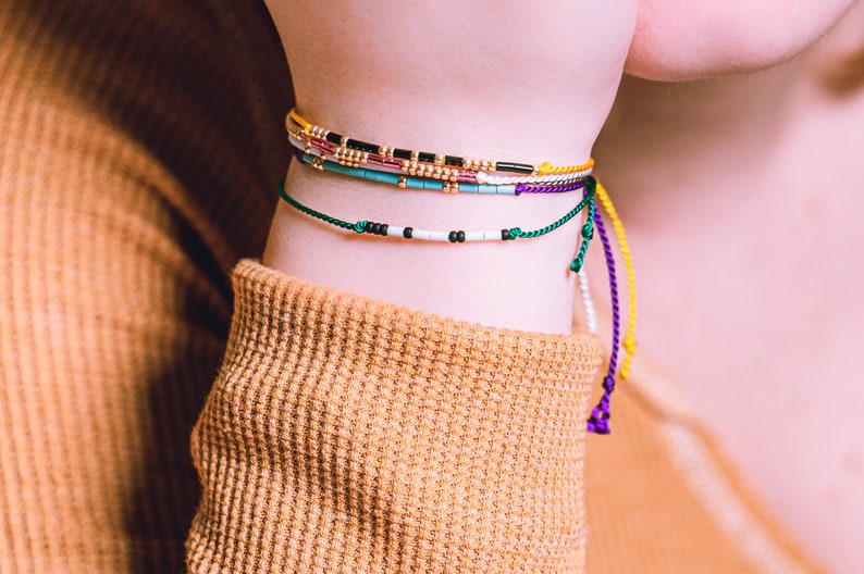 May include: A close-up of a person's wrist wearing several colorful beaded bracelets. The bracelets are made of small beads in various colors, including green, blue, yellow, purple, and black. The bracelets are tied with knots and have long, dangling ends.