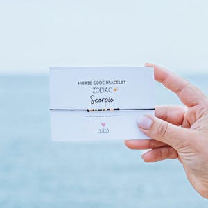 Peut inclure: Un bracelet en cordon noir avec des perles dorées et noires qui épellent "Scorpion" en code Morse. Le bracelet est sur une carte blanche avec le texte "Bracelet en code Morse Zodiac Scorpion" et un petit cœur rose avec le mot "POPSY".