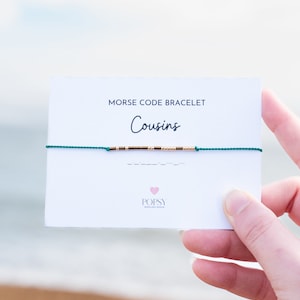 May include: A green string bracelet with gold beads spelling out "Cousins" in Morse code on a white card with the text "Morse Code Bracelet" and the brand name "POPSY".