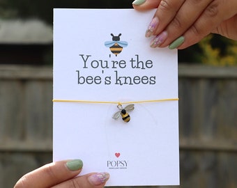 May include: A white card with the text "You're the bee's knees" and a small bee illustration. A yellow string is attached to the card, with a bee charm hanging from it. The charm is silver and yellow. The card is held by a person.