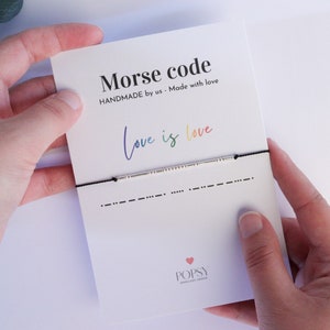 May include: A black string bracelet with a silver bar charm that says "Love is Love" in Morse code. The bracelet is on a white card with the text "Morse code" and "HANDMADE by us - Made with love".