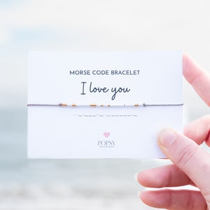 May include: A white card with the text "Morse Code Bracelet I love you" and a grey string bracelet with gold beads. The bracelet is spelled out in Morse code. The card also has the text "POPSY Jewellery Design" with a pink heart.
