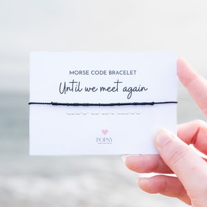 May include: A white card with black text that reads "Morse Code Bracelet Until We Meet Again" and a black string bracelet with black beads. The bracelet is in Morse code. The card has a pink heart with the word "POPSY" in black text.